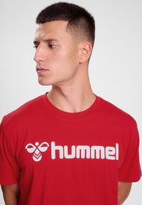 Rød bomulds t-shirt med et stort hvidt "hummel" logo og et bi-grafik. Korte ærmer, rund halsudskæring, glat tekstur.