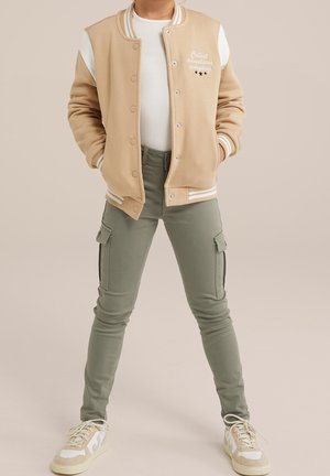 Person iført beige varsityjakke med teksten "Collect adventures everyday", hvid skjorte, grønne cargobukser og beige sneakers, stående med hænderne i lommerne.