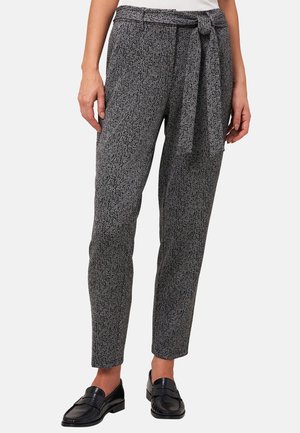 Frau trägt hochtaillierte, grau gemusterte, konisch zulaufende Hose mit Taillenschärpe, kombiniert mit schwarzen Slip-On-Loafers, steht vor weißem Hintergrund.