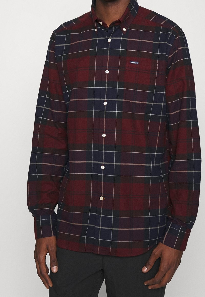 Homme portant une chemise à carreaux bordeaux et bleu marine, avec une petite poche poitrine et des boutons blancs, debout devant un fond uni.