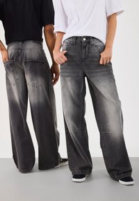 Dos personas vistiendo jeans de mezclilla gris deslavado de pierna ancha. Los pantalones tienen cinco bolsillos y una textura descolorida que pasa de oscuro a claro.