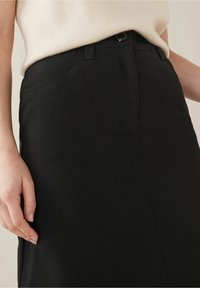Detailní záběr ruky ženy odpočívající na boku černé sukně, v kombinaci se světle zbarveným tričkem s krátkým rukávem.