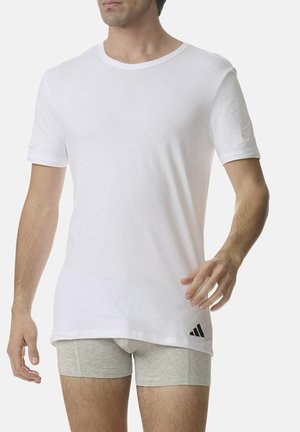 Weißes T-Shirt mit kurzen Ärmeln aus Baumwolle, mit rundem Halsausschnitt und einem schwarzen Adidas-Logo nahe dem Saum. Kombiniert mit grauen Shorts.
