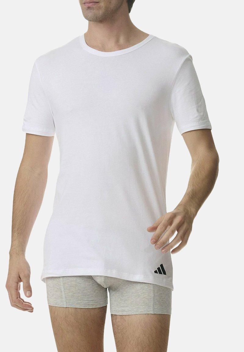 T-shirt bianco a maniche corte in cotone, con scollo tondo e logo Adidas nero vicino all'orlo. Abbinato a pantaloni corti grigi.