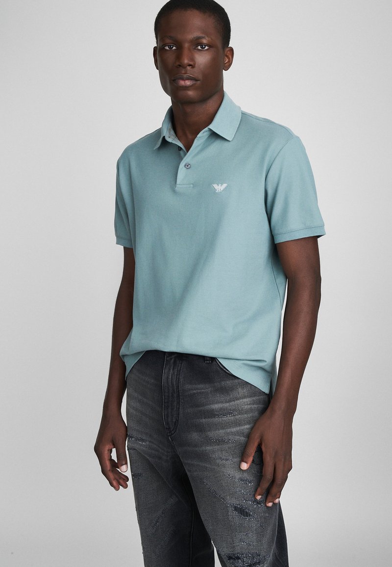 Polo bleu clair avec col, présentant un petit logo brodé sur la poitrine. Associé à un jean foncé déchiré.