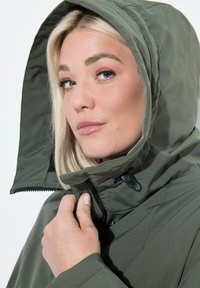 Blondynka w zielonej kurtce z kapturem, trzymająca suwak blisko szyi, patrząca lekko w bok z neutralnym wyrazem twarzy.