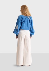 Blusa in cotone blu con maniche a pallone e ricami floreali bianchi; abbinata a pantaloni bianchi a gamba larga; tessuto morbido e testurizzato.