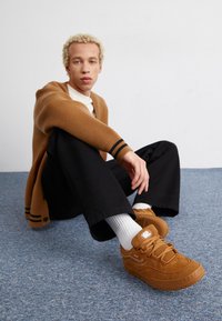 Braune Wildleder-Sneaker mit weißen Akzenten, flachen Schnürsenkeln und Logopatch. Das Modell trägt einen cremefarbenen Rollkragenpullover, eine braune Strickjacke und schwarze Hosen.