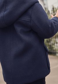 Marineblauer Fleece-Jacke mit Kapuze, die eine weiche Textur, Fronttaschen und eine lockere Passform aufweist. Details zeigen die Stoffnaht und die Platzierung der Taschen.