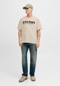 Beżowy, oversize t-shirt z grafiką "STUDIO Jack & Jones", noszony z niebieskimi dżinsami i beżowymi, casualowymi butami, dopełniony jasnoszarym czapką.