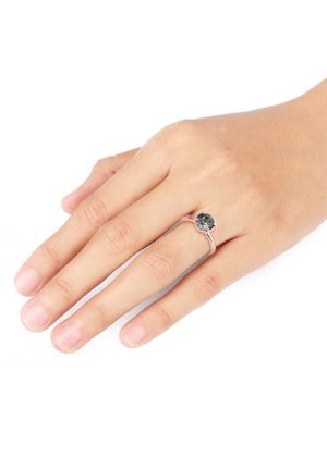 Rosaguld ring med en rund, mørk sten på en slank ring, holdt på en hånd med naturlige negle mod en hvid baggrund.