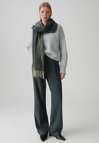 Maglione grigio peloso, sciarpa verde scuro con frange, pantaloni a vita alta verde scuro e scarpe nere a punta. Sfondo semplice.
