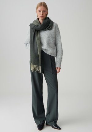 Grijze fuzzy trui, donkergroene sjaal met franjes, high-waisted donkergroene broek en zwarte puntige schoenen. Eenvoudige achtergrond.