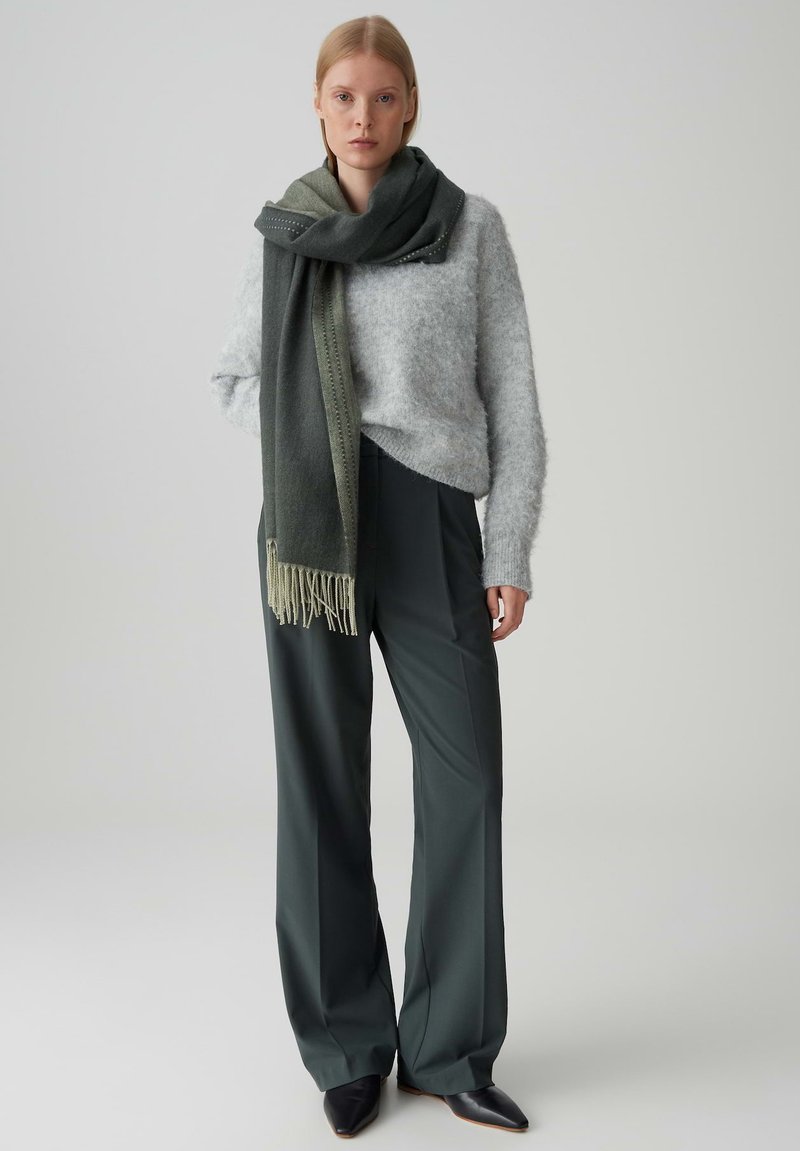 Maglione grigio peloso, sciarpa verde scuro con frange, pantaloni a vita alta verde scuro e scarpe nere a punta. Sfondo semplice.