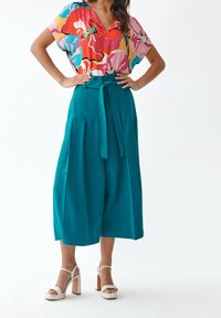 Kolorowa kwiatowa bluzka z głębokim dekoltem w kształcie litery V, zestawiona z wysokim stanem, miętowymi, szerokimi spodniami z wiązanym paskiem oraz białymi sandałami na słupku.