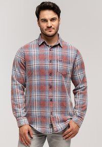 Mężczyzna w czerwonej, szarej i białej koszuli w kratkę z guzikami i kieszonką na piersi, stojący na tle jasnego, jednolitego tła.