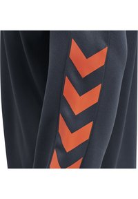 Tissu sombre avec quatre flèches chevrons rouges audacieuses disposées verticalement sur une manche ou une jambe de pantalon.