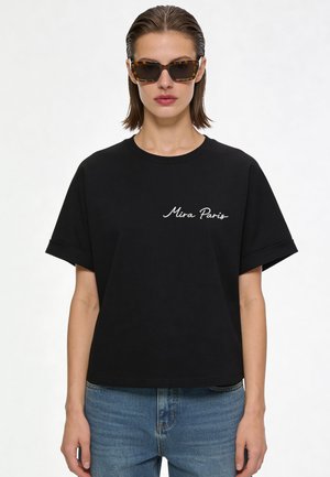 Femme aux cheveux courts et mouillés portant des lunettes de soleil en écaille, un t-shirt noir "Mira Paris" et un jean bleu, debout sur un fond blanc.