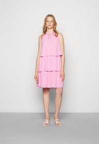 Vestido rosa em camadas com um tecido texturado, apresentando um fecho de pescoço com laço e um design fluido. Usado com sandálias brancas de tiras.