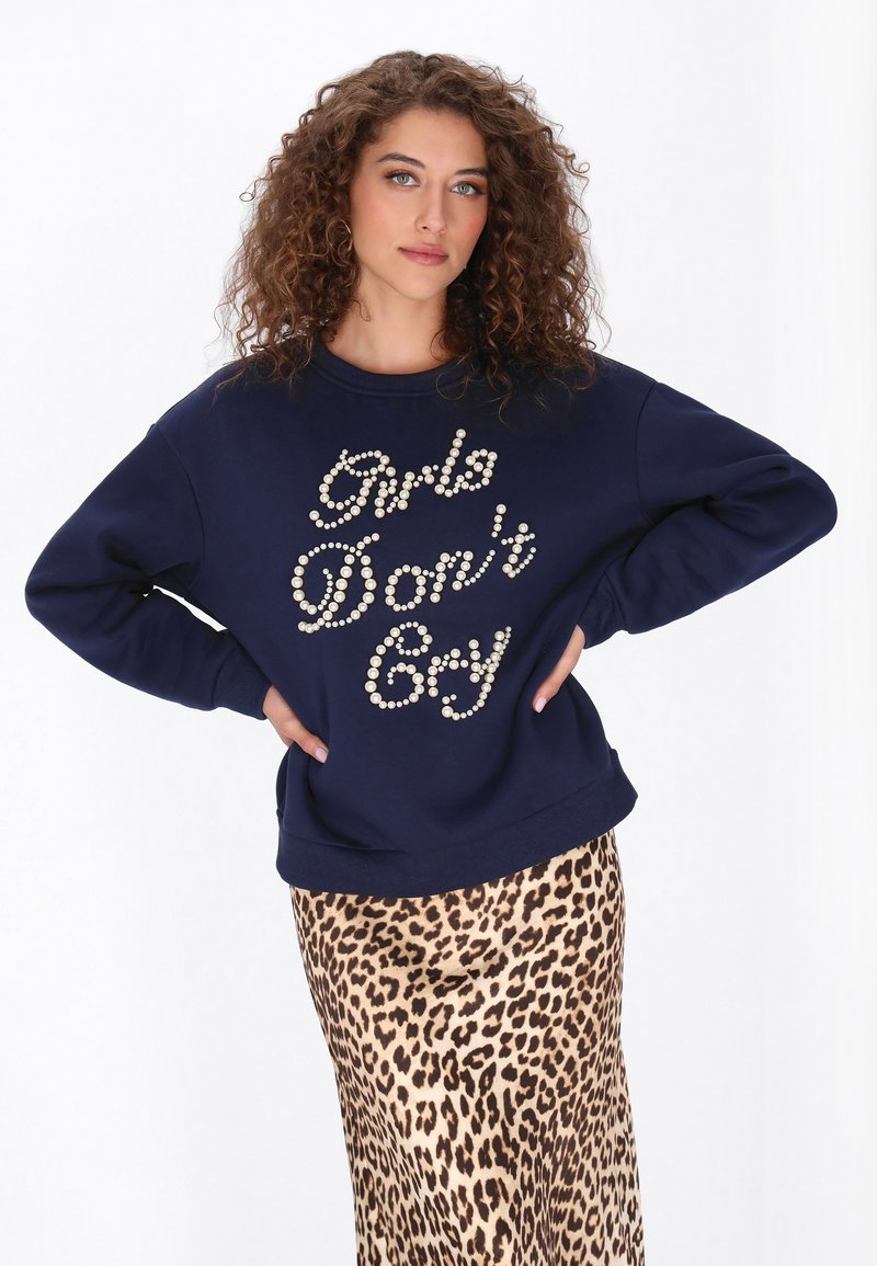 Donkerblauwe trui met 'Girls Don't Cry' in parelaccenten. Gecombineerd met een satijnen rok met luipaardprint. Krullend haar, handen in de zij.
