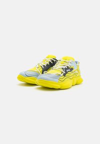 Sportiga sneakers med en design i gult, grått och ljusblått. Texturerad tygöverdel, tjock gult sula och intrikata vågmönster. Vita snören.