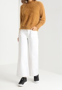 Maglione marrone peloso, jeans bianchi a gamba larga e sneakers nere con suole bianche. Tessuto strutturato e vestibilità comoda in un outfit casual.