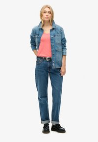 Kvinde stående iført en denimjakke over en koralfarvet t-shirt, blå jeans med sort bælte og sorte snørestøvler mod en hvid baggrund.