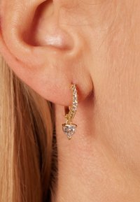 Boucle d'oreille en or avec un pendentif en forme de cœur en pierre précieuse transparente, ornée de petites pierres scintillantes le long de la boucle. Texture lisse.