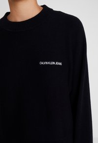 Czarny dziany sweter z okrągłym dekoltem i małym białym wyszywanym logo "CALVIN KLEIN JEANS" na przodzie. Miękka faktura i luźny krój.