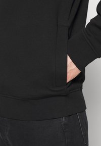 Mustat hupparit, jossa on ribbattu helma ja hihansuut sekä sivutasku. Kangas näyttää pehmeältä ja tekstuuriltaan miellyttävältä, sopii rentoon käyttöön.