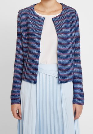 Vrouw draagt een blauwe, roze en witte getextureerde open-front jas over een wit topje en een lichtblauwe geplooide rok met een riem.