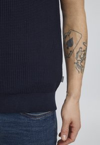 Marineblå teksturert strikkegenser med ribbestrikket kant. Har en sideetikett og en nærbilde av en tatovert arm som holder stoffet.
