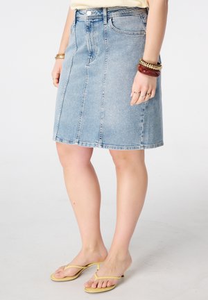 Femme portant une jupe en jean bleu clair, des sandales à talons jaunes et des bracelets, vue de la taille aux pieds sur un fond uni.