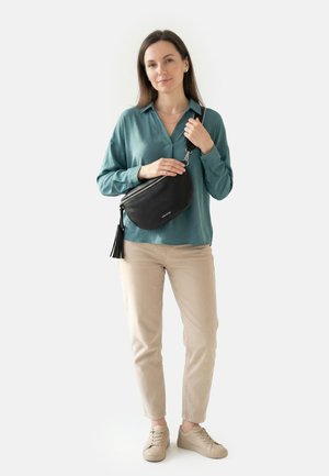 Frau trägt eine türkisfarbene Bluse, beige Hosen und beige Sneakers, hält eine schwarze Leder-Umhängetasche mit Quasten-Detail.