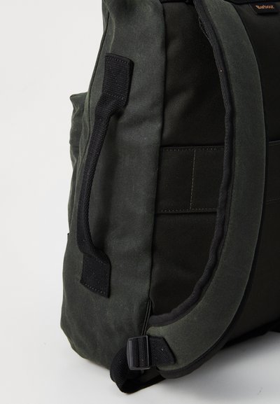 Vue de côté d'un sac à dos vert foncé montrant une sangle rembourrée, une poignée en tissu noir et des détails de couture sur fond blanc.