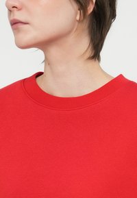 Oseba s kratkimi temnimi lasmi, oblečena v svetlo rdečo majico s krožnim izrezom, prikazana od vratu do sredine prsi v stranskem pogledu.