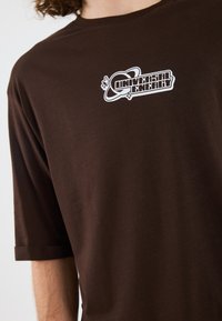 Camiseta de manga corta marrón hecha de tejido suave que presenta un logotipo gráfico blanco que dice "Energía Universal" con un diseño de planeta.