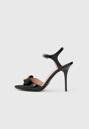 Sandales à talons hauts en cuir verni noir avec une bride de cheville, bout ouvert, accent nœud et détails en métal doré. Texture lisse et design épuré.