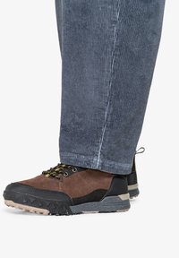 Bruine suède en zwart synthetisch laagste schoen met een gestructureerde zool, gouden accenten en zwarte veters; gecombineerd met grijze corduroy broek.