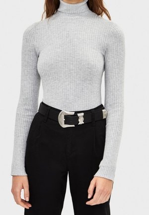 Femme portant un pull à col roulé côtelé gris clair rentré dans un pantalon noir avec une ceinture noire munie d'une boucle argentée.