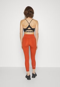 Reggiseno sportivo nero con schiena incrociata, abbinato a leggings arancioni a vita alta e aderenti e scarpe da ginnastica nere.