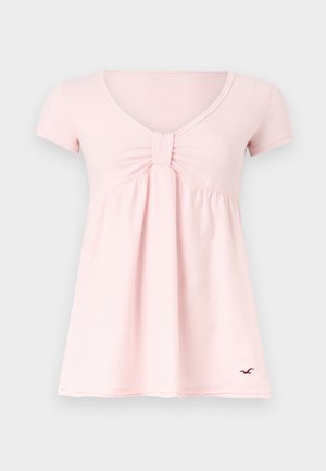 Haut pour femmes rose clair à manches courtes et col en V, avec un détail noué froncé au niveau de la poitrine et un petit logo oiseau noir près de l'ourlet.