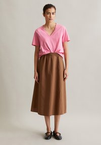 Rosa V-Ausschnitt T-Shirt kombiniert mit einem braunen Maxirock mit elastischem Bund aus strukturiertem Stoff; das Model trägt schwarze flache Schuhe.