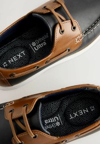 Chaussures en cuir noir et marron à lacets avec des semelles blanches. L'intérieur présente une semelle intérieure à motif, portant l'inscription "Ortholite Ultra."