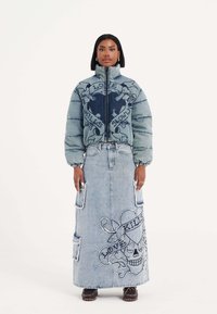 Jeans pufferjakke med lynlås foran, prydet med et blåt og gråt grafikdesign; parret med en lang jeansnederdel, der viser et broderet kranium.