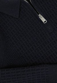 Tamno plavi dukserica s patentnim zatvaračem i teksturiranim uzorkom, s visokim ovratnikom i metalnim patentnim zatvaračem. Tkanina izgleda mekano i pletiva.