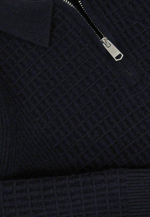 Tumši zili rāvējslēdzēja džemperis ar teksturētu rakstu, kuram ir augsta apkakle un metalizēts rāvējslēdzējs. Audums izskatās mīksts un adīts.