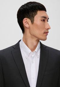 Schwarzes Blazer mit Reverskragen über einem weißen Hemd. Glatte Textur, taillierte Passform, Seitenansicht, die die Gesichtszüge zeigt.