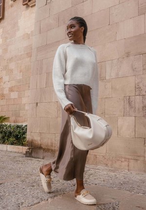 Light gray knitted sweater, brown satin skirt, cream bag, and beige shoes with gold accents; set against a stone wall with plants.