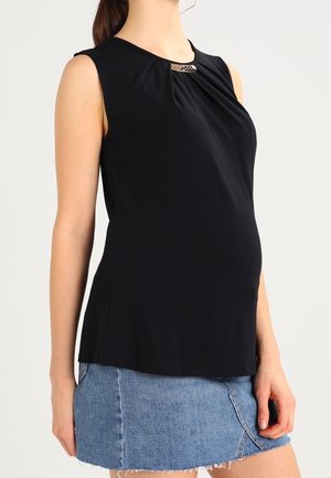 Kvinde iført ærmeløs sort top med dekorativ spænde og blå denim mini nederdel mod ensfarvet baggrund.